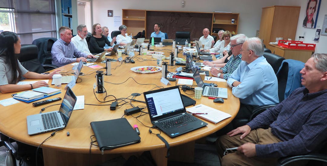 A photo of 14 people sitting around a large conference table with papers and laptop computers in front of them.