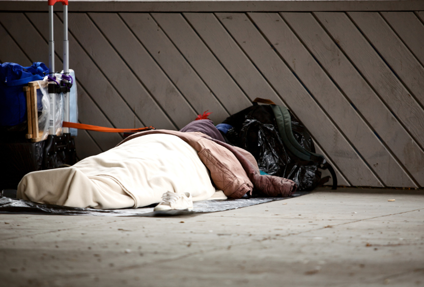A photo of a person sleeping on the ground outside. They are covered in blankets and surrounded by luggage and other bags.